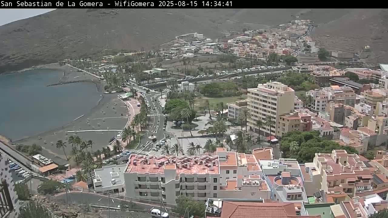 An elevated view shows a coastal town with a dark-sand beach and calm water, under a mostly clear sky.