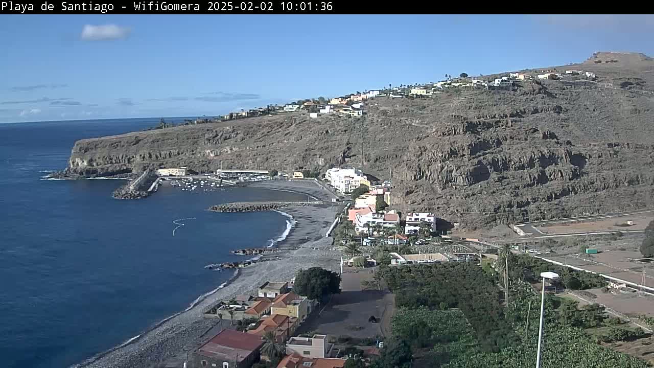 A rocky, coastal village with a small harbor and pebble beach is visible on a clear, sunny day.