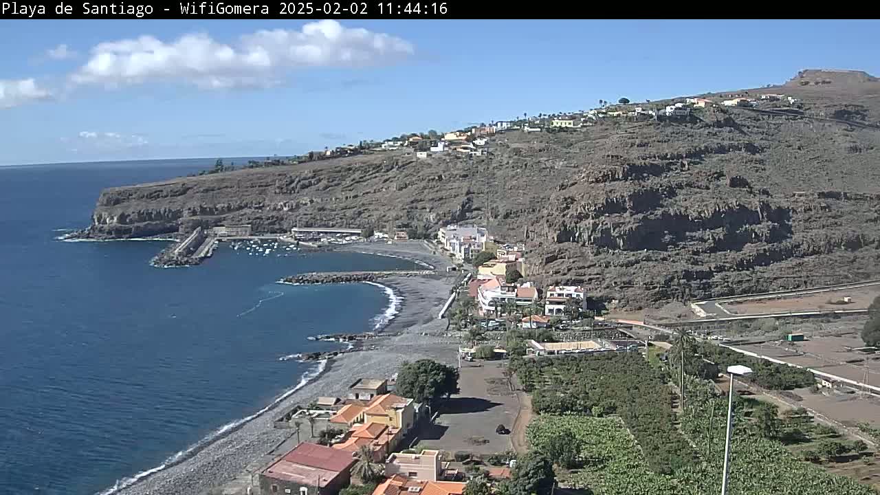 A mostly sunny day reveals a rocky, dark-colored hillside overlooking a small, dark-sand beach and a calm bay with a few boats.