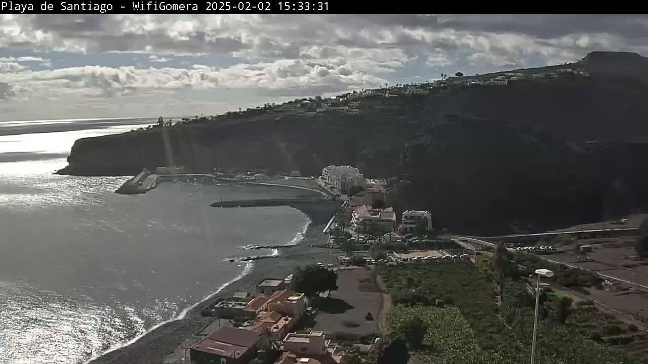 A coastal town with a dark-sand beach sits below a dark, rocky cliff under a partly cloudy sky.