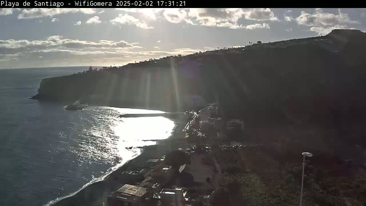 A partly sunny day shows a dark, rocky cliff overlooking a beach and dark-blue ocean; the sun's rays are visible on the water.