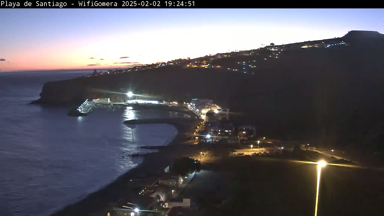 An aerial nighttime view of a coastal town, with the last light of twilight fading into the dark, shows a dark-colored beach, a small harbor, and buildings illuminated on a hillside overlooking the ocean.