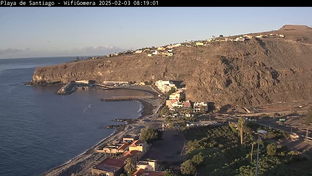 A dark-sand beach curves along a rocky coastline under a clear, bright sky, with buildings and vegetation nestled along the shore and up a steep, arid hillside.