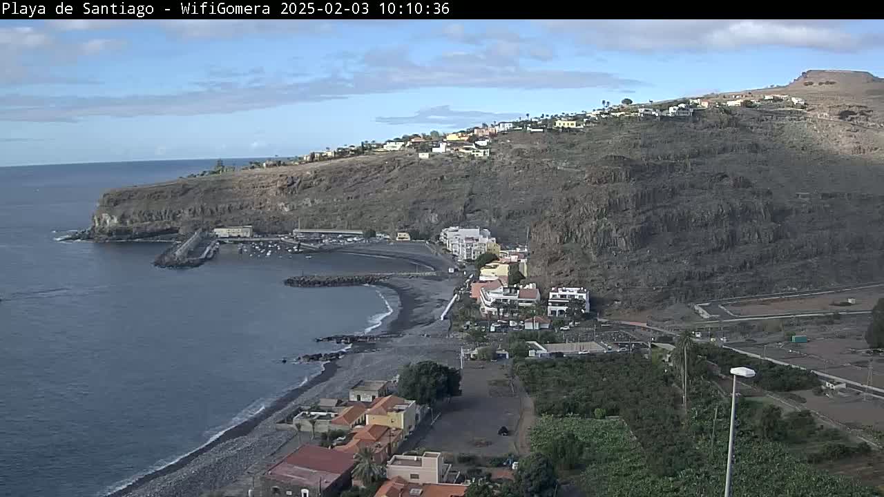 A mostly sunny day reveals a coastal town nestled against a dark, rocky cliffside, with a small, dark-sand beach and a calm ocean.
