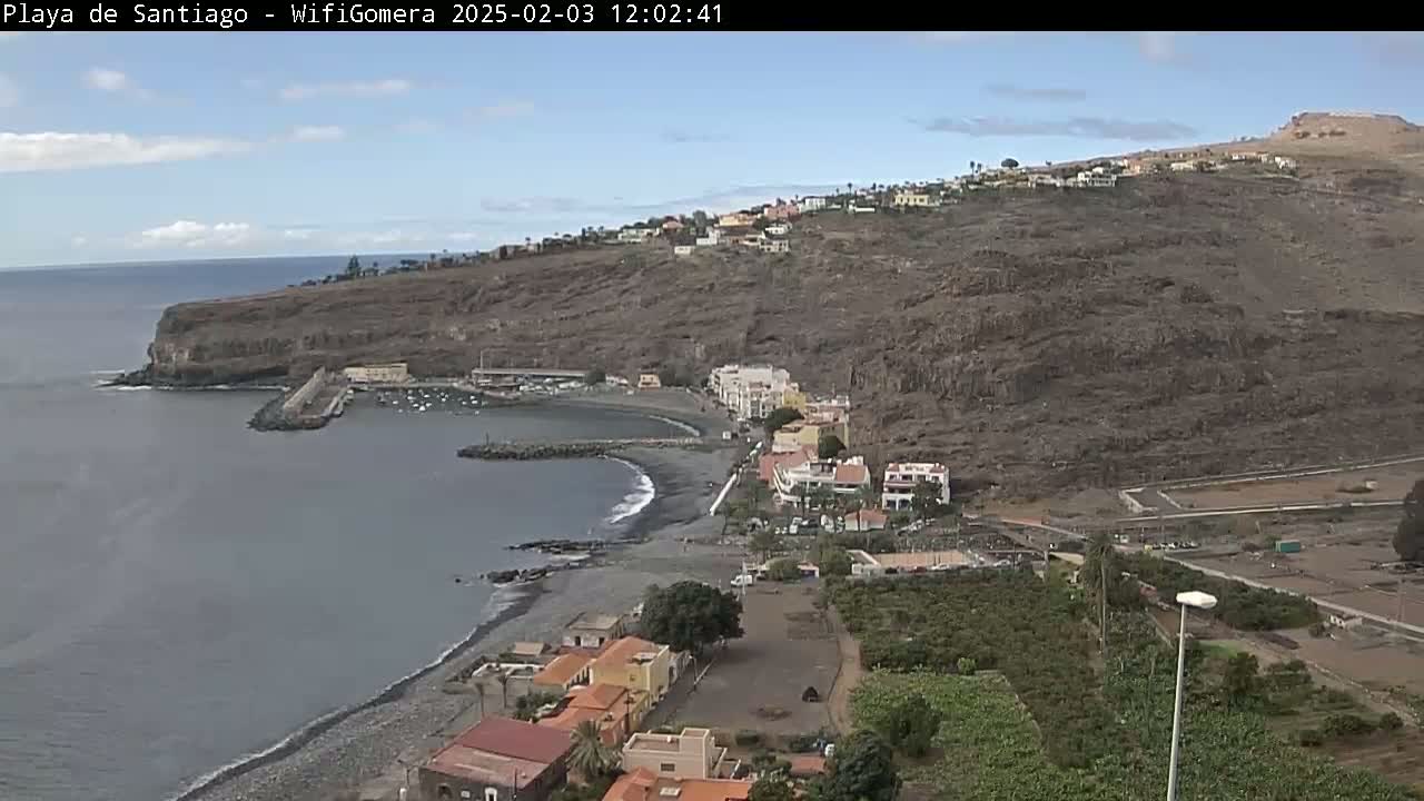 A coastal town nestled against a steep, dark-colored hillside, overlooking a calm, gray ocean under a mostly clear sky.