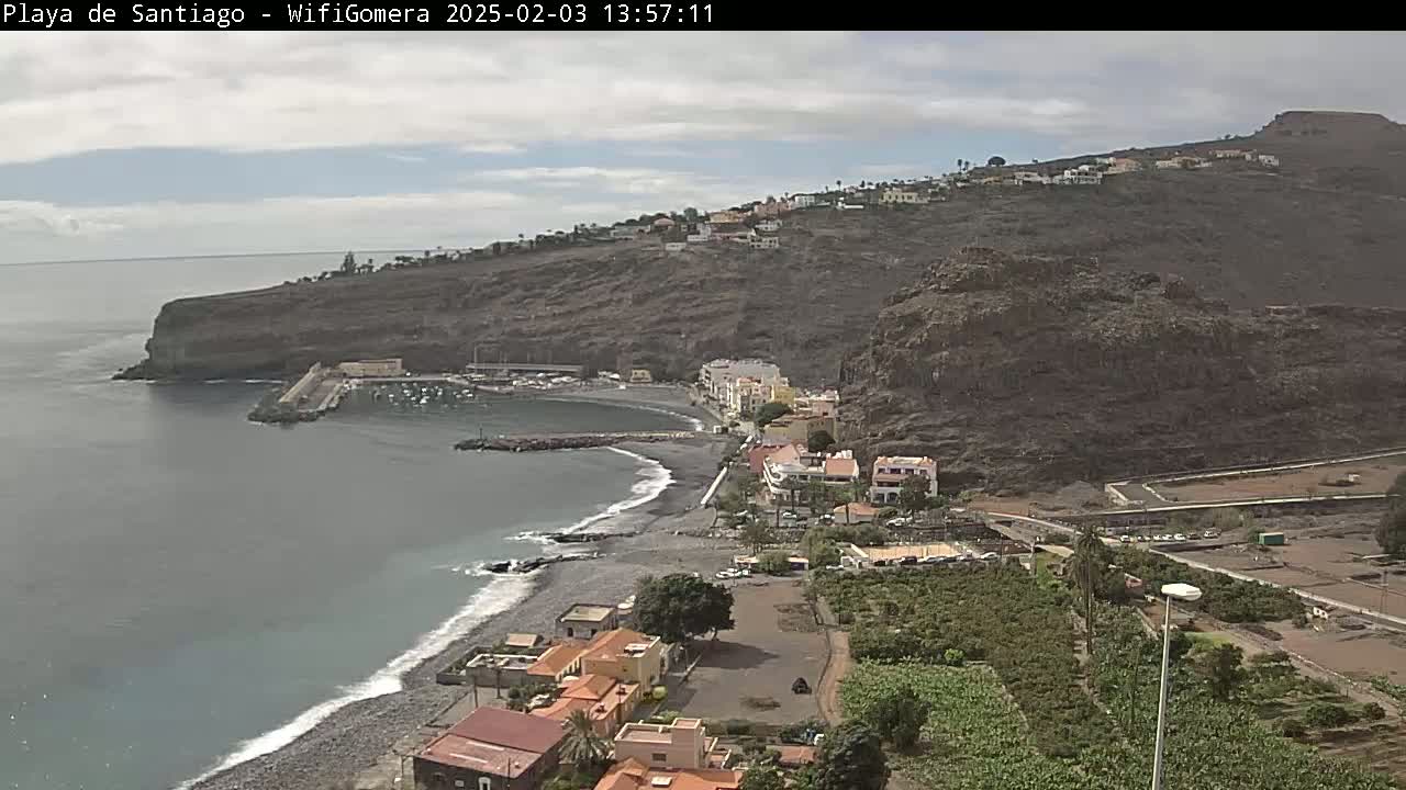 A partly cloudy day shows a black sand beach curving into a small harbor next to a village nestled at the base of a dark, rocky cliff.