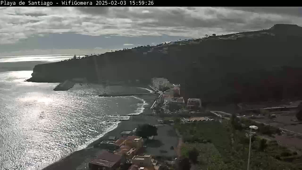 A mostly cloudy day overlooks a dark-sand beach and coastal town nestled against a steep, dark hillside, with a calm ocean reflecting sunlight.