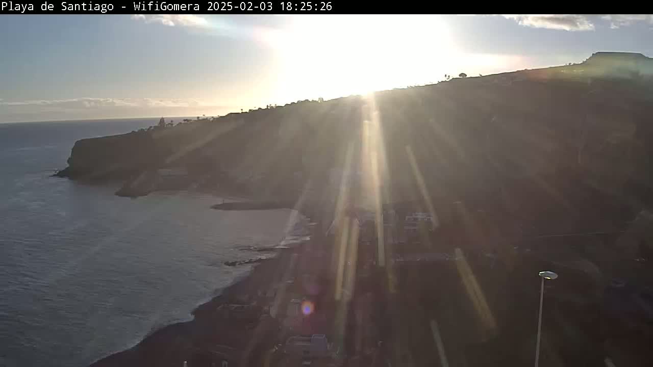A coastal scene at sunset shows a dark, rocky shoreline and ocean with bright sun rays shining through the atmosphere.