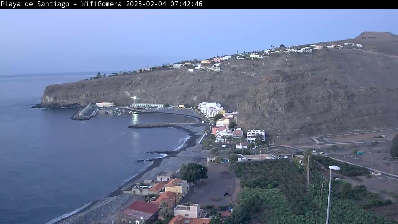 A coastal town nestled against a dark, rocky cliff overlooking a calm, dark-blue ocean under a twilight sky.