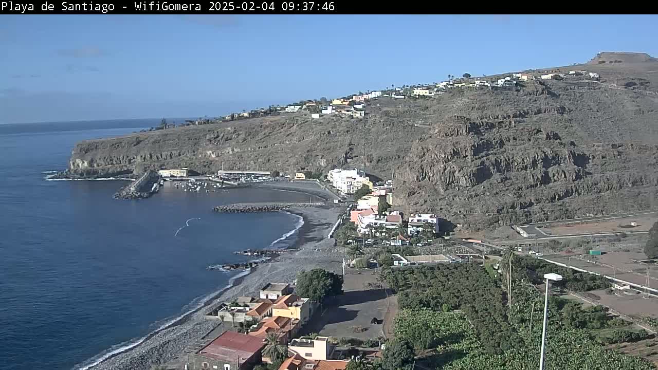 A dark-colored, sandy beach curves along a rocky coastline next to a small town nestled on a steep, arid hillside under a clear blue sky.
