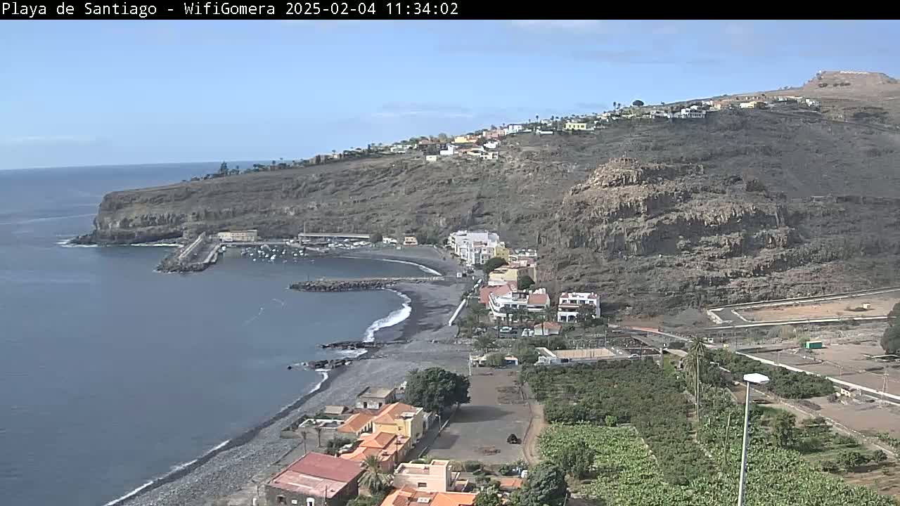 A coastal town nestled against a rocky hillside overlooks a dark-sand beach and calm ocean under a clear sky.