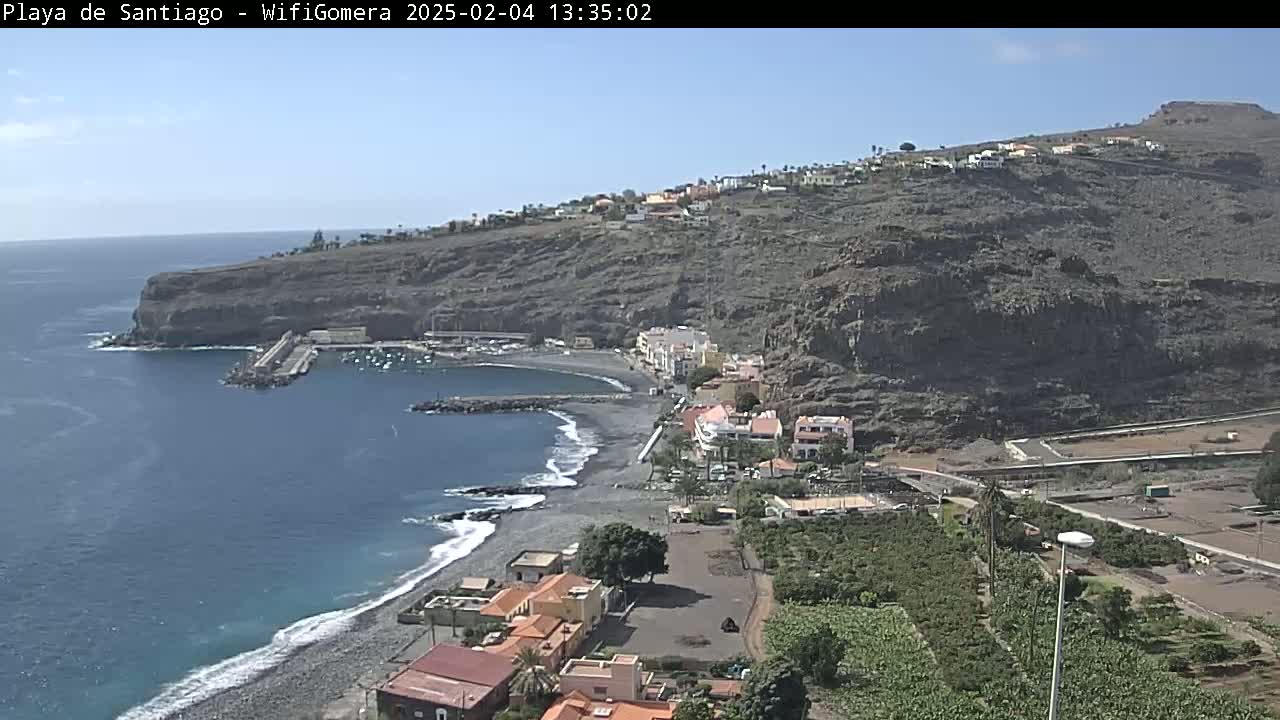 A coastal town nestled against a dark, rocky hillside overlooks a calm, blue ocean with a small, dark-sand beach and a harbor.