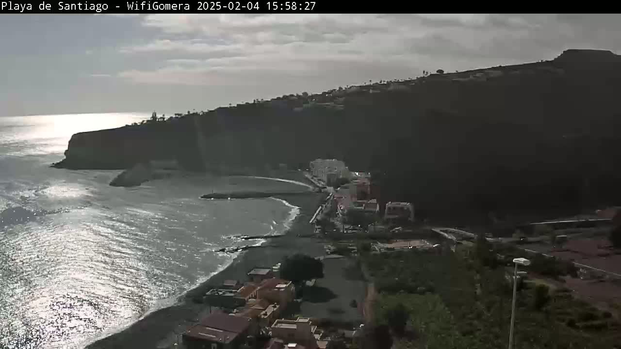 A coastal town sits nestled against a dark, mountainous hillside, with a calm, gray ocean reflecting sunlight.