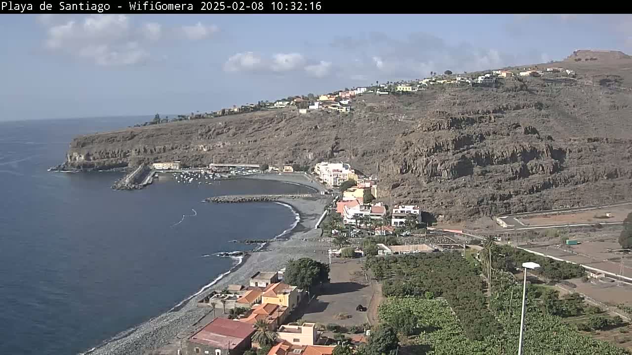 A mostly sunny day shows a rocky coastline with a small town and harbor nestled against a dark, steep hillside, overlooking a dark blue ocean.