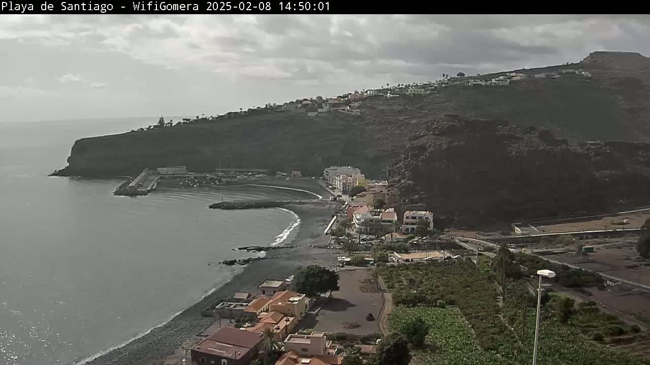 A coastal town nestled against a dark volcanic cliff overlooks a dark-sand beach and calm ocean under an overcast sky.