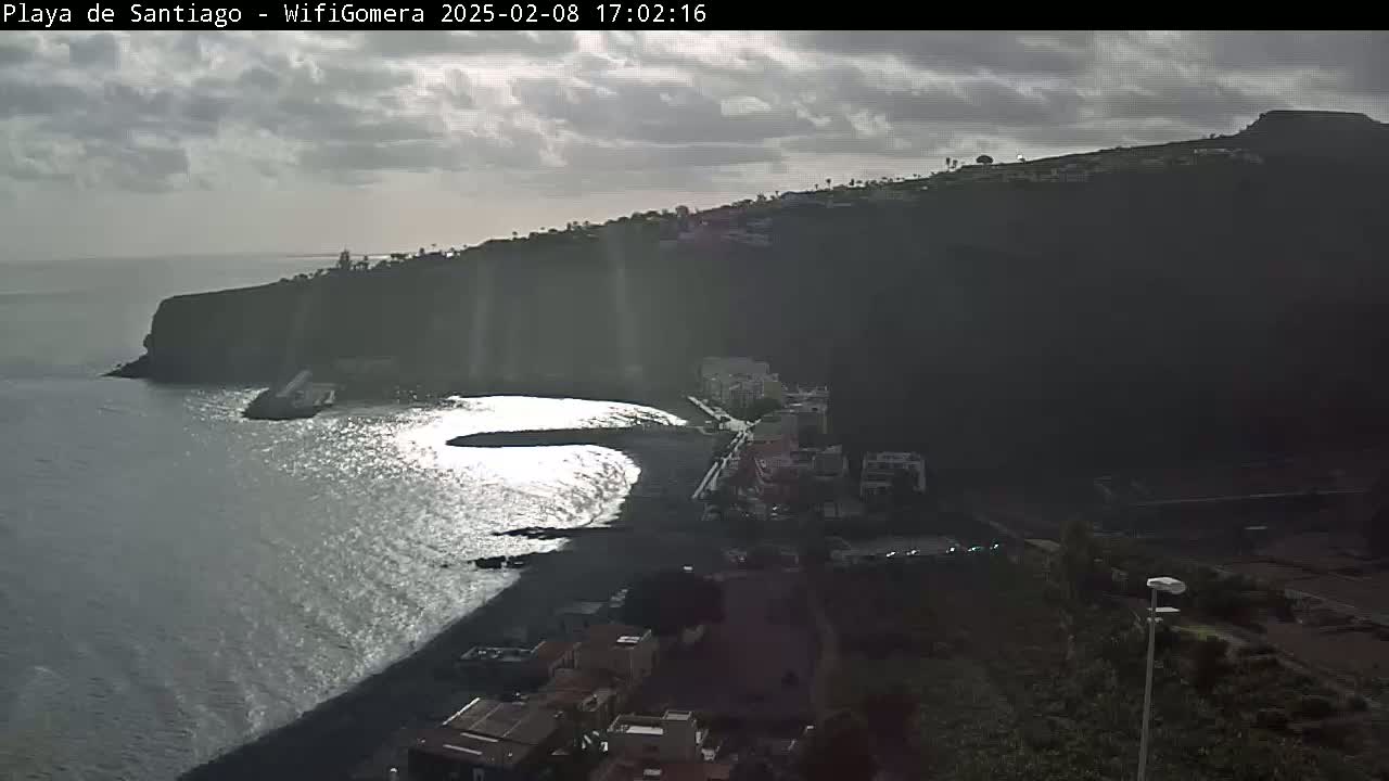 A dark, partly cloudy day overlooks a black sand beach nestled below a steep cliffside, with buildings lining the shore and the ocean gently lapping at the sand.