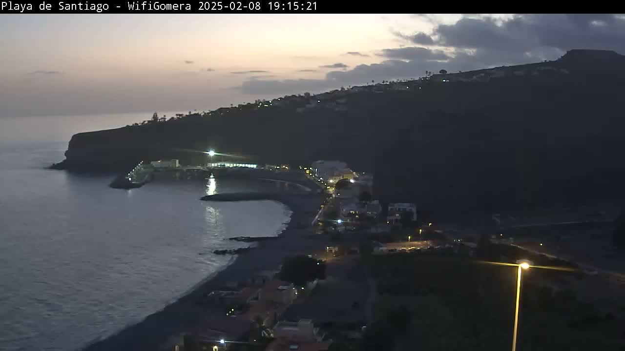 A dark, twilight view shows a coastal town nestled against a dark mountainside, overlooking a calm, dark ocean.