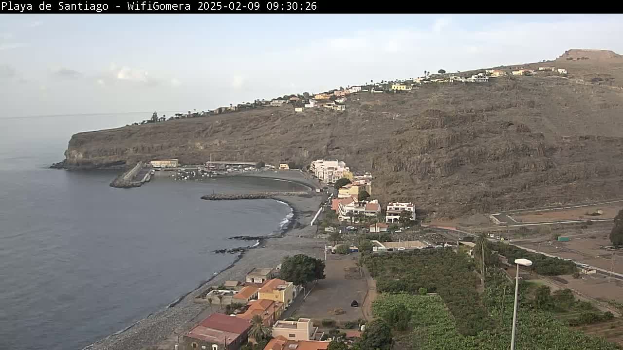 A coastal town nestled against a rocky hillside overlooks a calm, dark-grey ocean under a mostly cloudy sky.