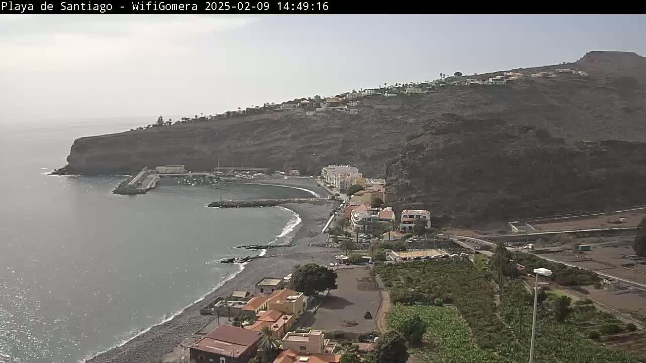 A mostly sunny day overlooks a dark-sand beach curving into a small harbor next to a dark volcanic cliffside village.