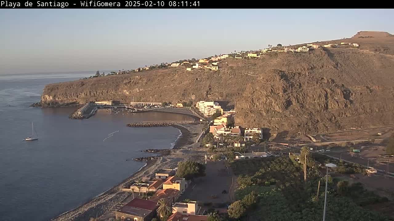 A coastal town nestled against a rocky, sunlit hillside overlooks a calm bay with a single sailboat.