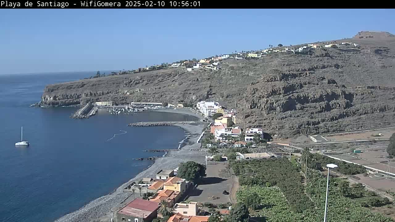 A coastal town nestled against a rocky hillside overlooks a calm, blue ocean harbor on a clear, sunny day.