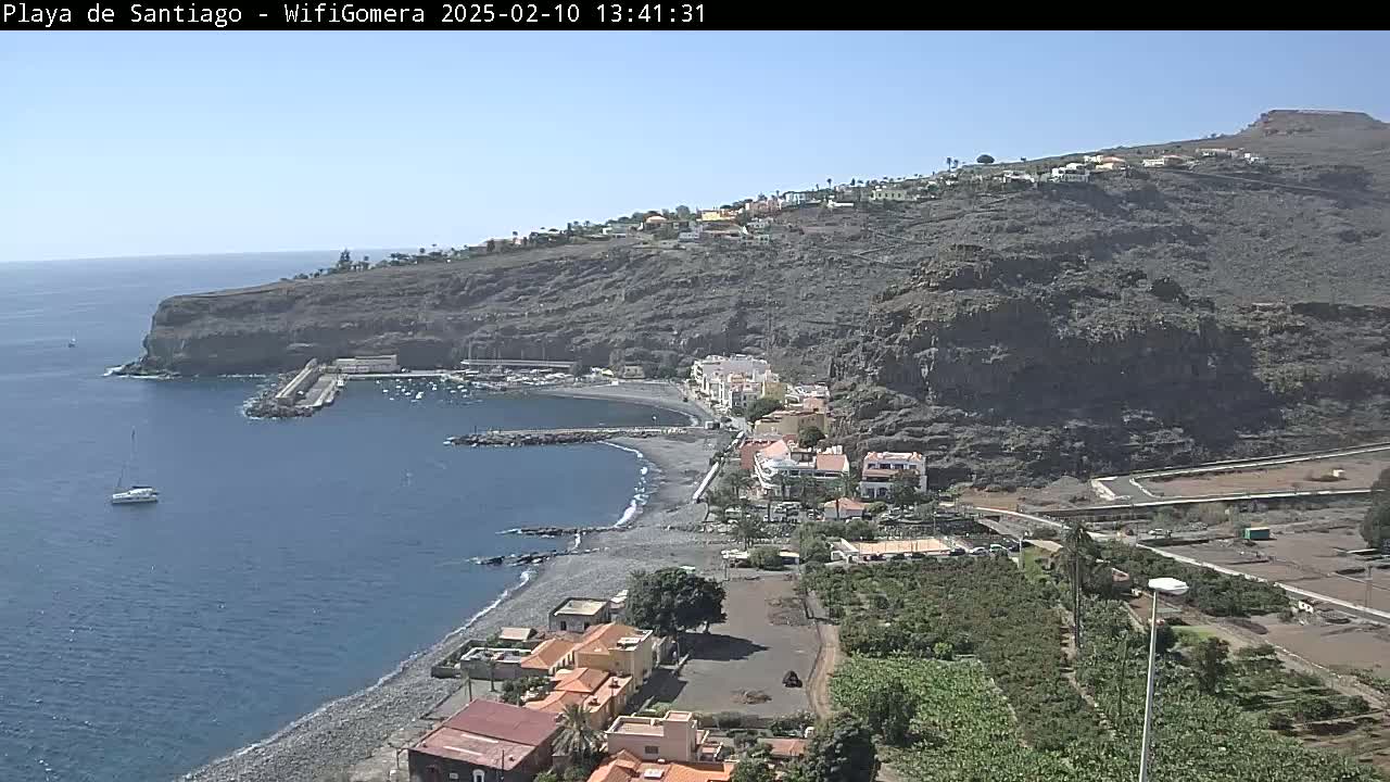 A coastal town nestled against a dark, rocky hillside, with a small harbor and a dark-sand beach, under a clear, sunny sky.