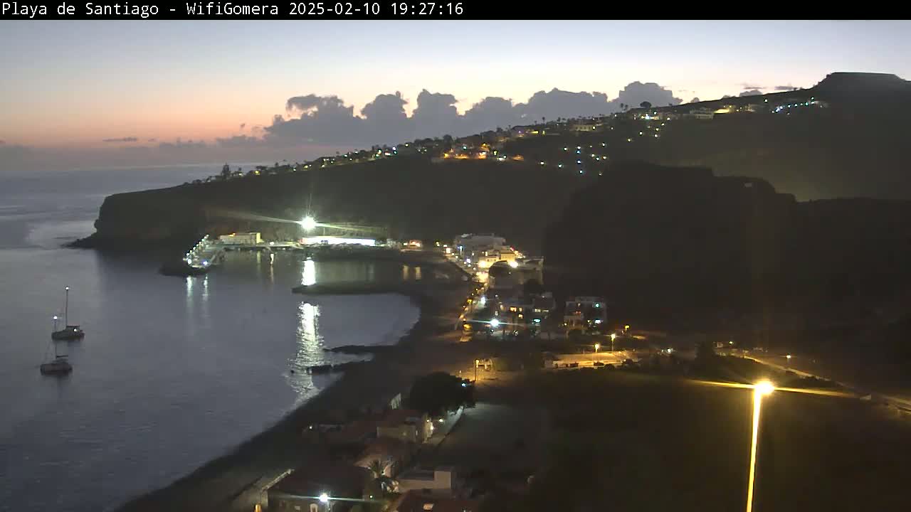 A nighttime coastal scene shows a dark hillside with illuminated buildings overlooking a calm bay where a few boats are moored, under a twilight sky with some clouds.