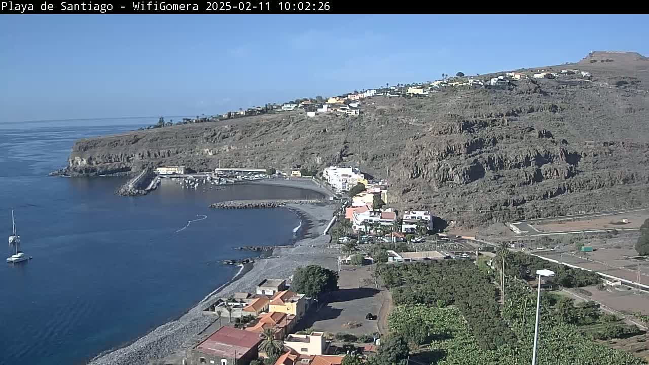 A coastal town nestled on a rocky hillside overlooking a calm bay with a small harbor and a pebble beach under a clear blue sky.