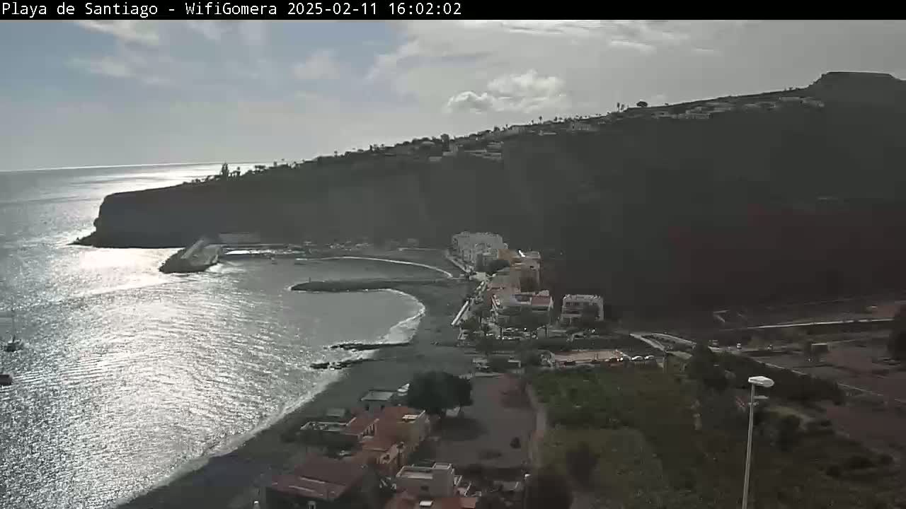 A partly cloudy day reveals a coastal town nestled against a dark cliff overlooking a calm, sunlit ocean.