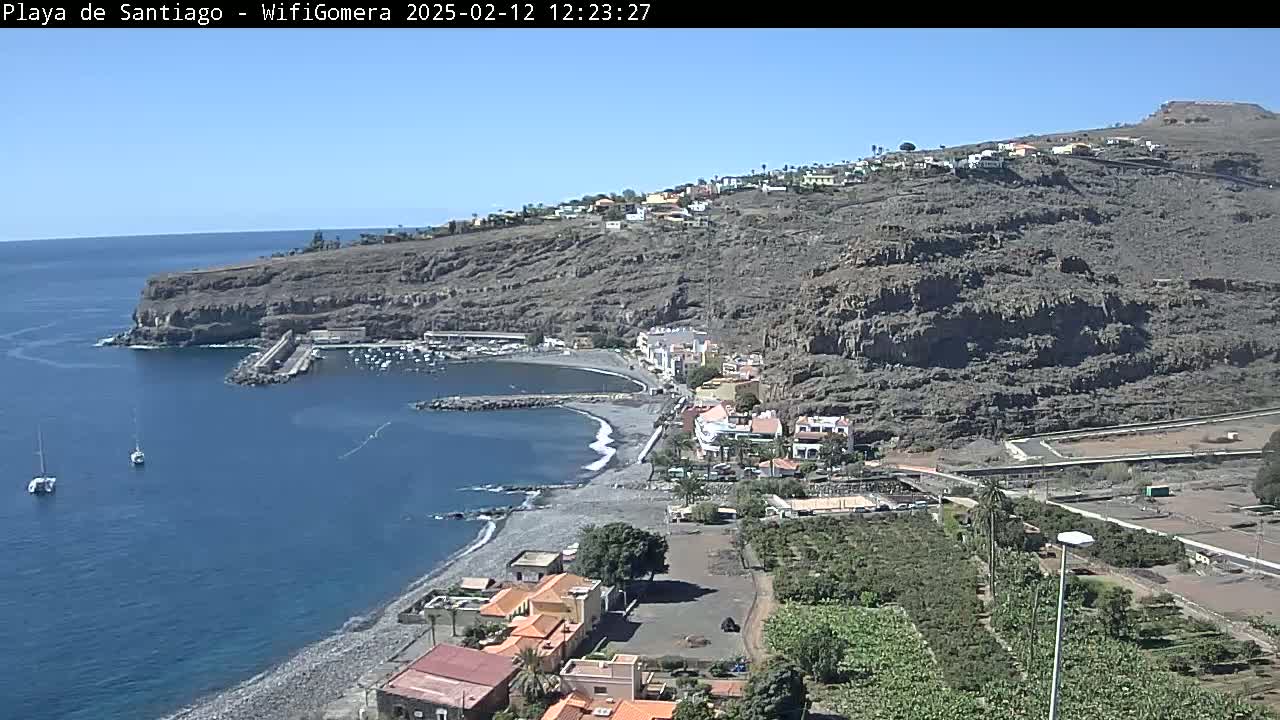 A coastal town with a small harbor nestled against a dark, rocky cliffside on a sunny day, overlooking a dark-sand beach and calm blue ocean with a few sailboats.