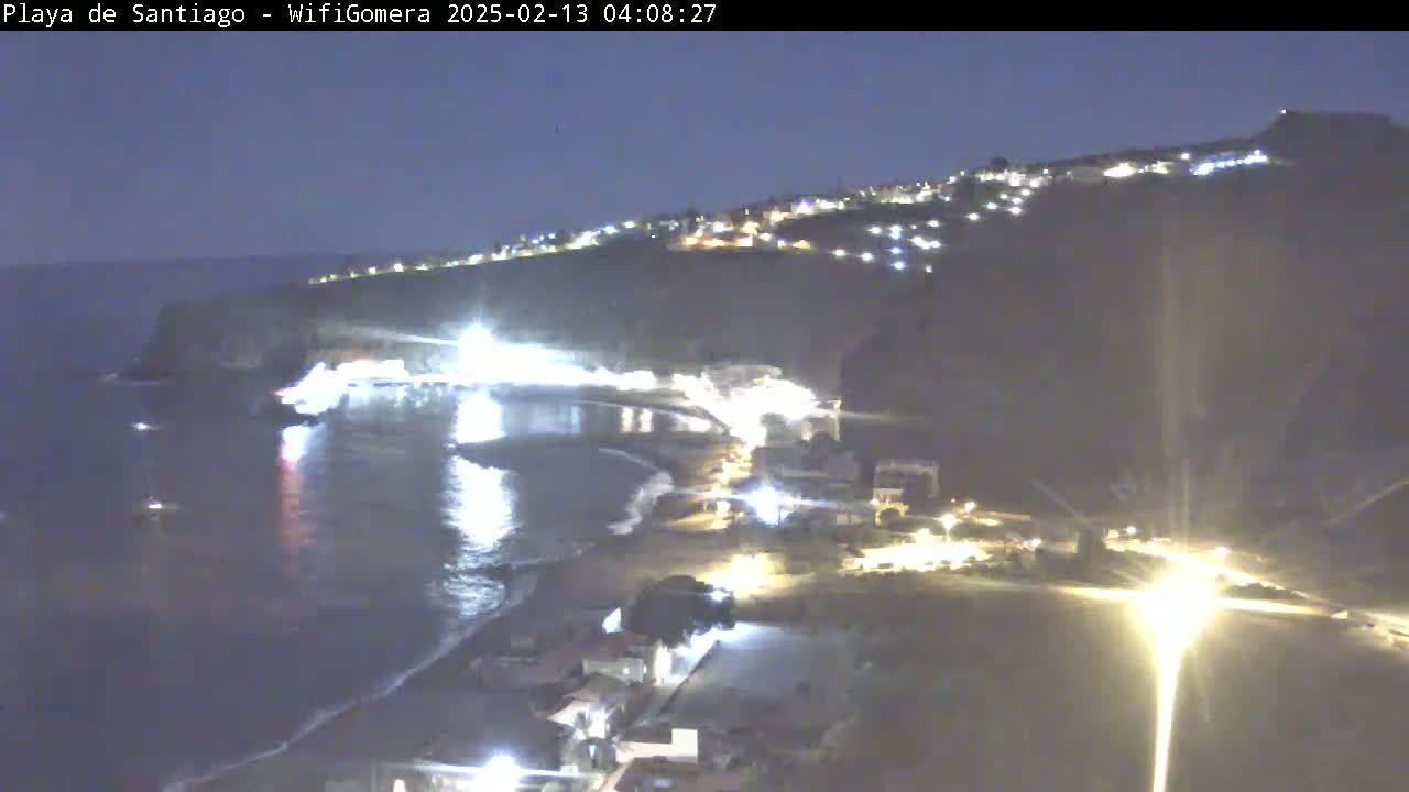 A nighttime, high-angle view shows a dark, rocky coastline with a beach and town illuminated by lights, overlooking a calm, dark ocean.