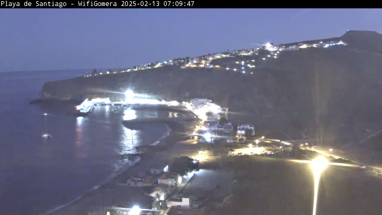 A nighttime coastal scene shows a dark-colored beach curving into a small bay, with a village of lights situated on a dark cliffside under a twilight sky.