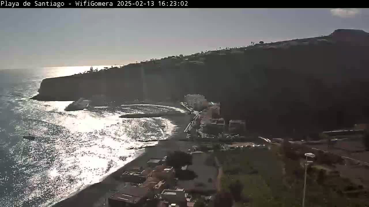 A partly sunny day shows a dark-sand beach meeting a rocky cliffside, with buildings and vegetation visible above and to the right of the beach, and gentle waves washing the shore.