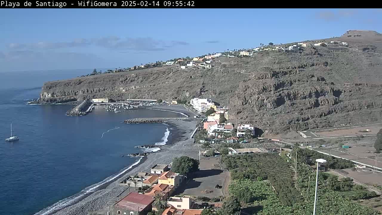A small, dark-sand beach and harbor are nestled below a hillside town, all under a clear, sunny sky.