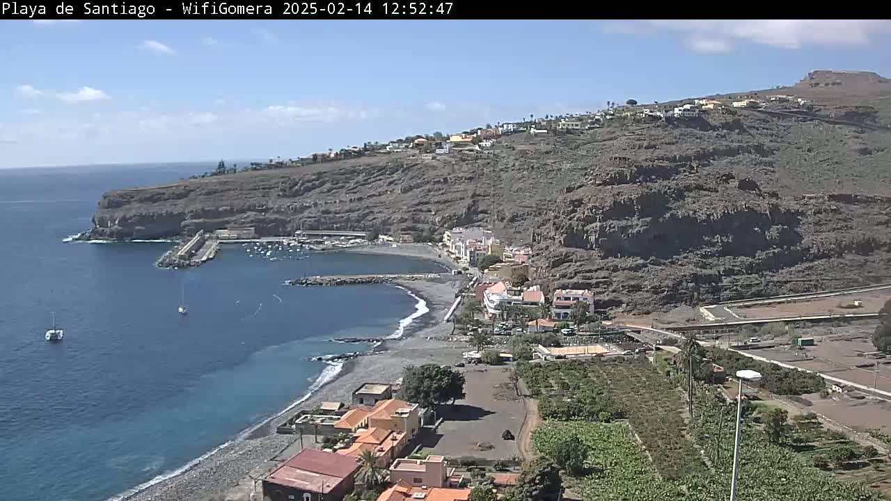 A coastal town nestled against a steep, rocky hillside overlooks a calm bay with several small boats, under a mostly sunny sky.