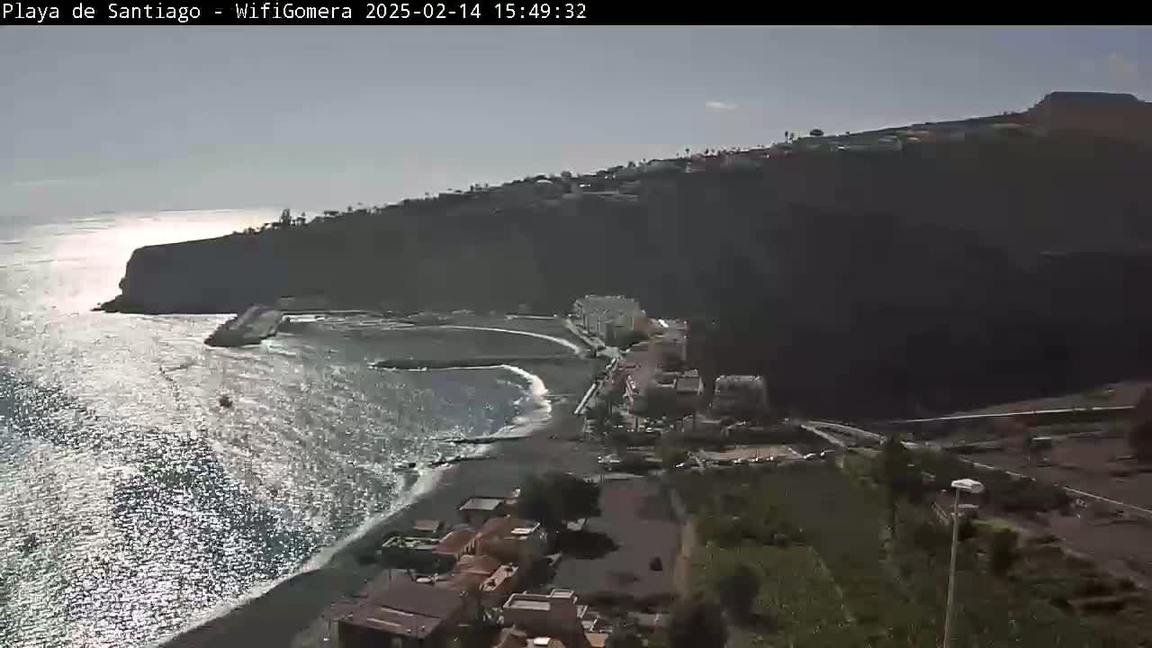A mostly sunny day overlooks a dark, rocky coastline with a small, dark-sand beach and a town nestled against the cliffside.