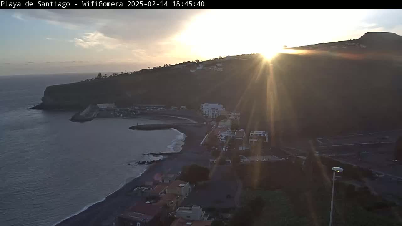 A coastal town with a dark sand beach is bathed in the bright light of a setting sun over a calm ocean.