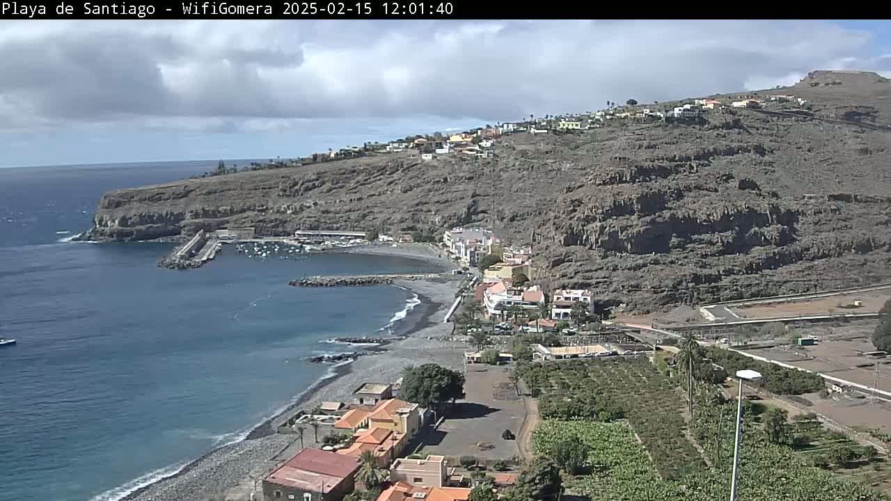 A mostly sunny day overlooks a dark-sand beach, small harbor, and coastal town nestled against a rocky hillside.