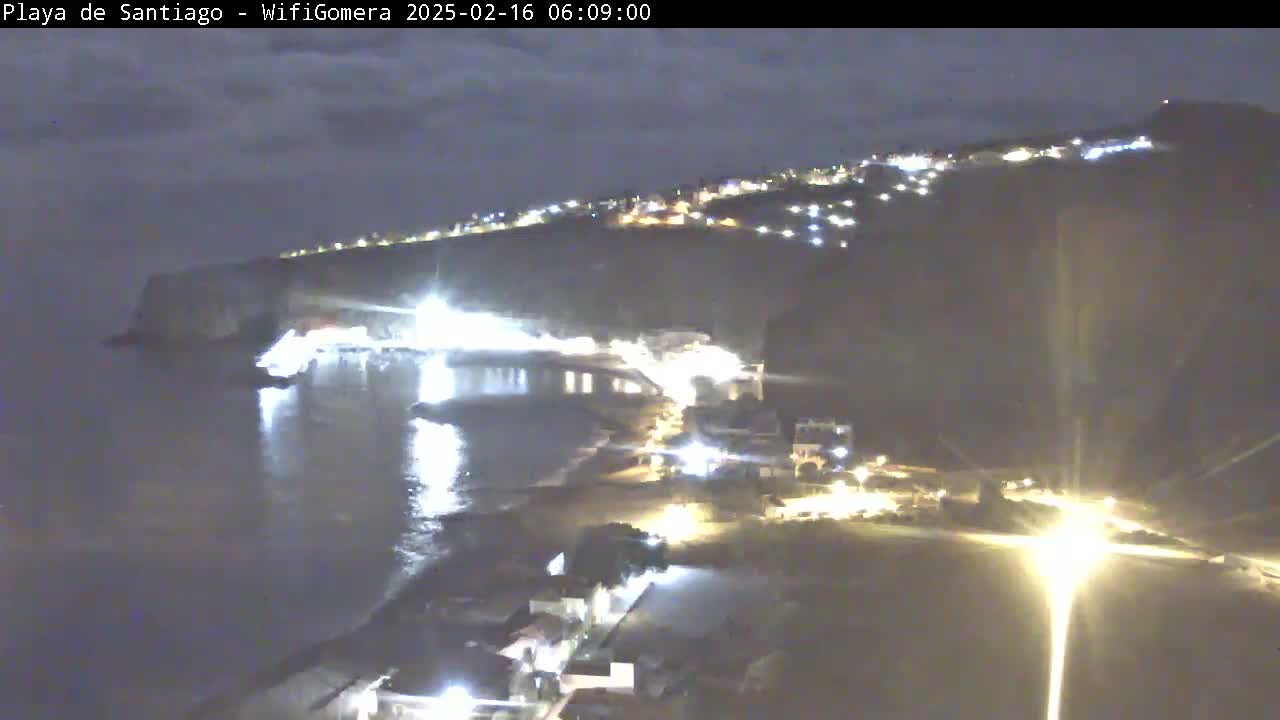 A nighttime coastal scene shows a dark, calm body of water reflecting lights from a town built into a cliffside.