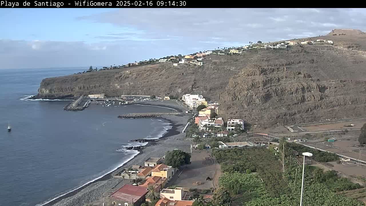 A partly cloudy day reveals a coastal town nestled against a dark, rocky hillside, with a small harbor and a dark-sand beach at the water's edge.
