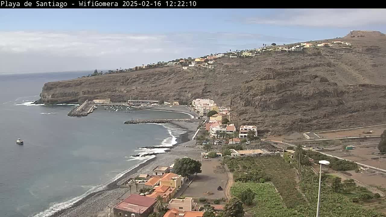 A coastal town nestled against a steep, rocky hillside overlooks a small harbor and dark-sand beach under partly cloudy skies.