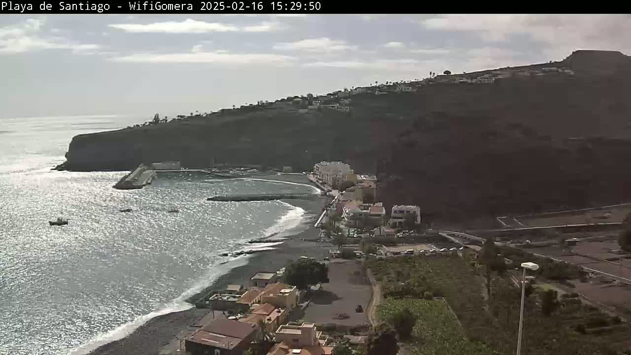 A mostly sunny day reveals a dark-sand beach and small town nestled below a dark, volcanic cliffside, next to a calm ocean with a few small boats.
