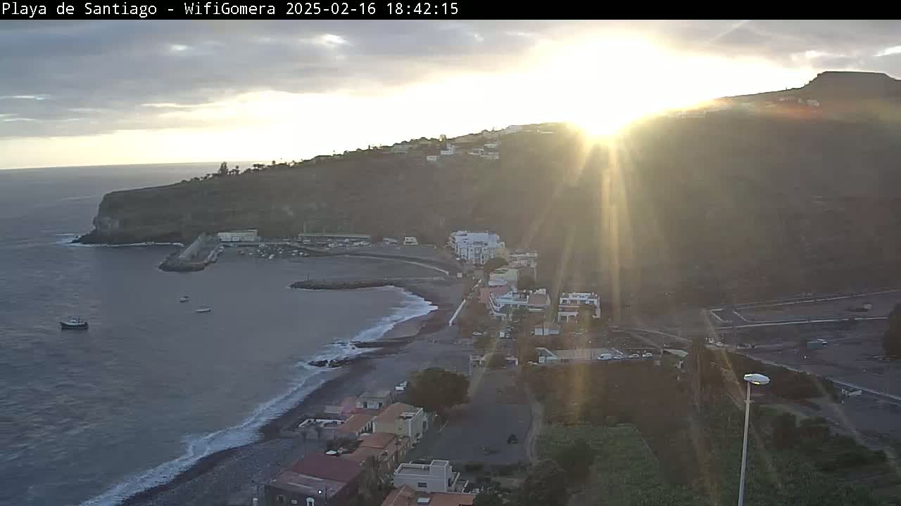 A coastal town nestled on a hillside, with a small harbor and dark sand beach, is bathed in the golden light of a setting sun under a mostly clear sky.