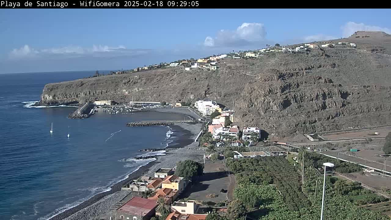A mostly sunny day overlooking a dark-sand beach and small coastal town nestled against a rocky hillside, with a calm ocean and a few sailboats visible.