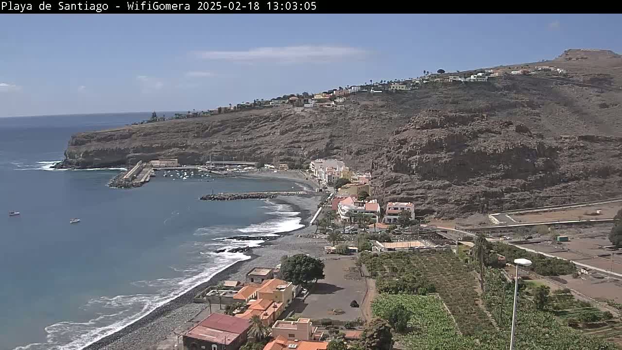 A black sand beach curves around a small harbor nestled below a rocky cliffside village under a mostly clear sky.