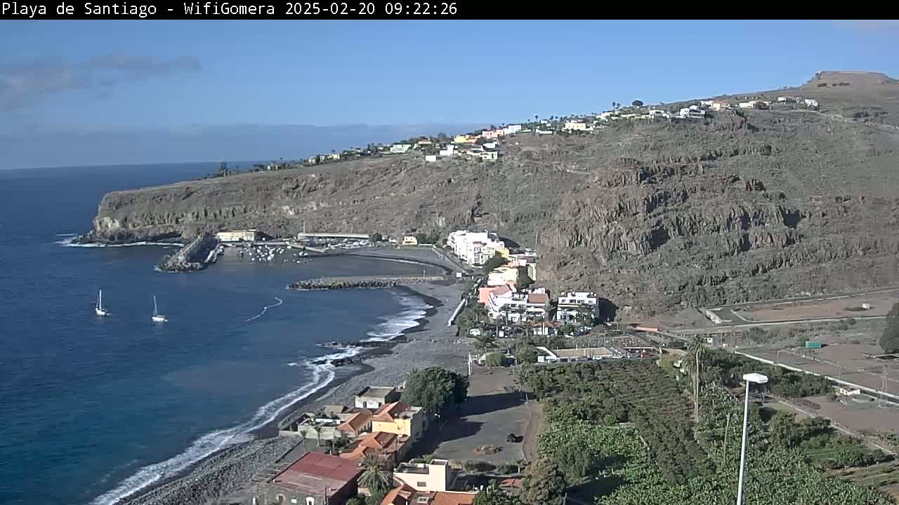 A coastal town with a dark-sand beach, a small harbor, and buildings nestled against a rocky hillside under a clear, sunny sky.