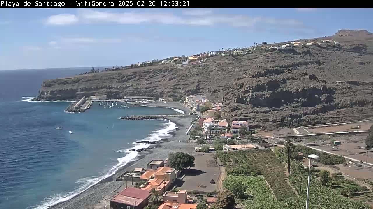 A dark-sand beach curves around a small harbor nestled between dark-colored cliffs under a mostly sunny sky.