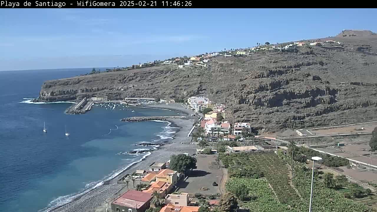 A coastal town nestled beside a dark-colored, pebble beach, with a small harbor and a rocky hillside rising behind it under sunny skies.