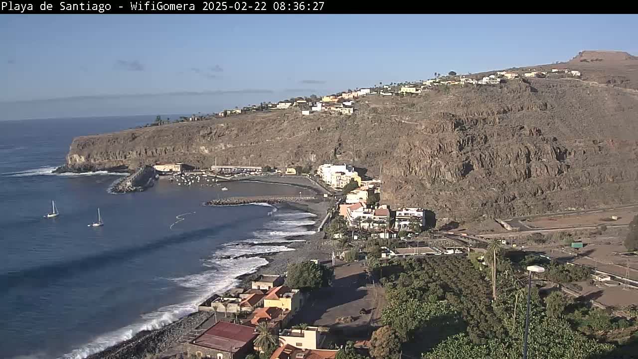 A coastal town nestled against a rocky hillside, with a small harbor and ocean waves breaking on a pebble beach under a clear sky.