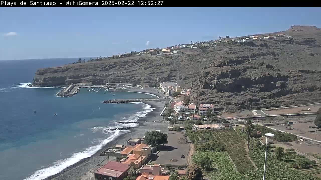 A dark-sand beach curves around a small harbor nestled below a rocky, arid hillside on a sunny day.